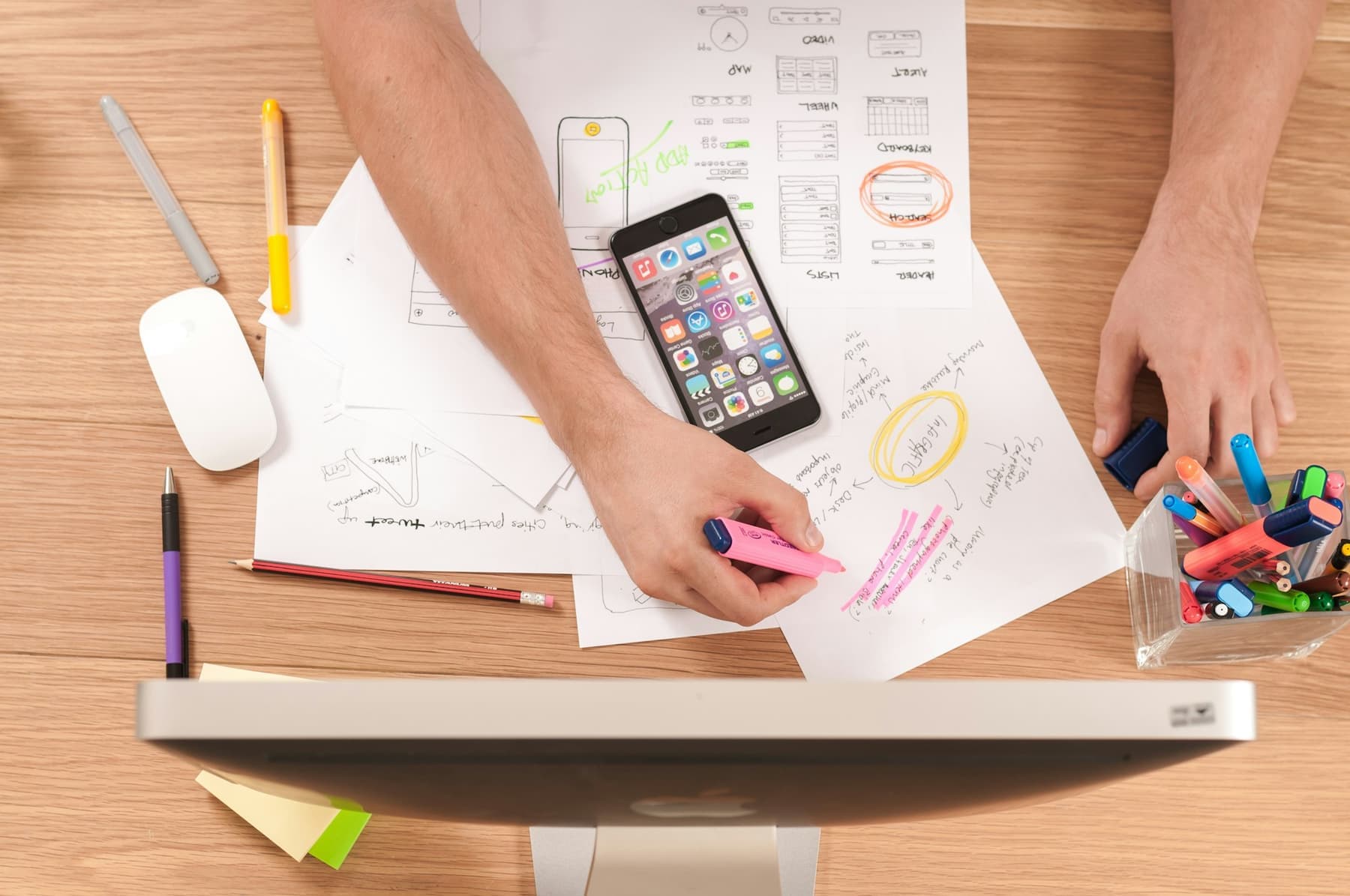 Top-down view of a designer sketching layouts with a phone and coloured pens on the desk.